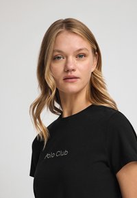 Camiseta negra de manga corta y cuello redondo. "Polo Club" está impreso en strass en la parte delantera. Textura de tela suave.