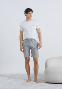 Balta īsroku T-krekls ar mazu logotipu, kombinēts ar gaiši pelēkām šortiem ar elastīgu jostasvietu un sānu kabatām. Basām kājām uz gaiša paklāja.