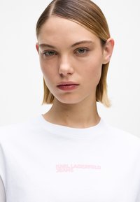 Valge puuvillane T-särk, mille rinnal on roosa "KARL LAGERFELD JEANS" logo. Ümar kaelus, sile tekstuur, klassikaline lõige.