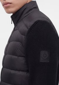 Gros plan d'une personne portant une veste matelassée noire avec des manches tricotées et un écusson logo Belstaff sur le bras gauche.