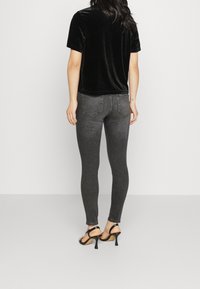 Mujer con blusa corta de terciopelo negro, jeans skinny gris oscuro y sandalias de tacón negro, de pie con la espalda hacia una pared lisa.