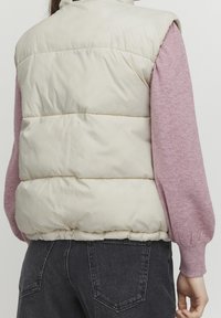 Beige vadderad väst med hög krage och horisontell quiltad design. Buren över en ljusrosa långärmad skjorta och mörkgrå jeans.