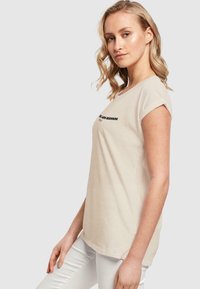 Beige t-skjorte med kort erme og rund hals, med svart tekst som sier "EN NY BEGYNDELSE." Myke bomullsmaterialer, avslappet passform og rullede mansjetter.