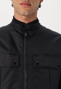 Černá bunda z hladké látky s vysokým límcem, předním zipem a dvěma náprsními kapsami se zapínáním na patentky.