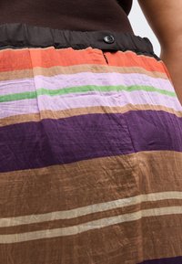 Πολύχρωμη φούστα με ρίγες σε πορτοκαλί, λεβάντα, πράσινο, μοβ και καφέ, με μαύρη ζώνη μέσης που κλείνει με κουμπί και φερμουάρ.