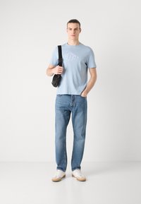 Lyseblå t-skjorte med kort erme og logo, kombinert med lyseblå rett-bente jeans og hvite sneakers med brune detaljer, bærende en svart crossover-veske.