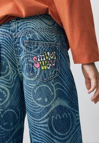 Jeans din denim cu un model albastru în spirală și un logo colorat „Smiley World” pe buzunarul din spate. Juxtapuse cu un top portocaliu.