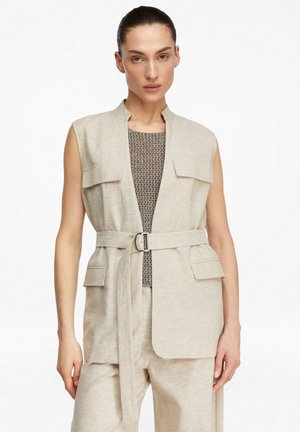 Frau trägt einen beigen ärmellosen Weste mit Gürtel über einem strukturierten grauen Top und passenden beigen Hosen, steht vor weißem Hintergrund.