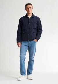 Navy work jacket in durable fabric, featuring a button-up front, two front pockets, and a chest pocket. Paired with light blue jeans and white shoes.