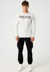 Camisa de manga larga, de color gris claro, con el texto negro "INSIDE" centrado en la parte delantera. Combinada con pantalones jogger negros y zapatillas blancas.