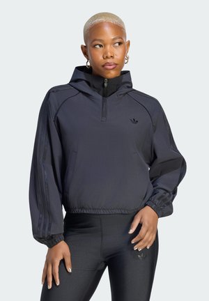 Osoba z krótkimi blond włosami, ubraną w czarną sportową kurtkę z kapturem oraz dopasowane czarne legginsy z logo Adidas na obu.