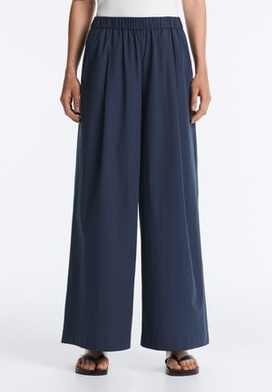 Personne portant un pantalon large bleu marine à taille élastique et des sandales noires, debout devant un fond blanc uni.