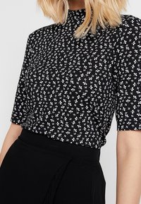 Top negro de manga corta con patrón floral blanco, cuello alto y tejido suave. Lleva un cinturón negro debajo.