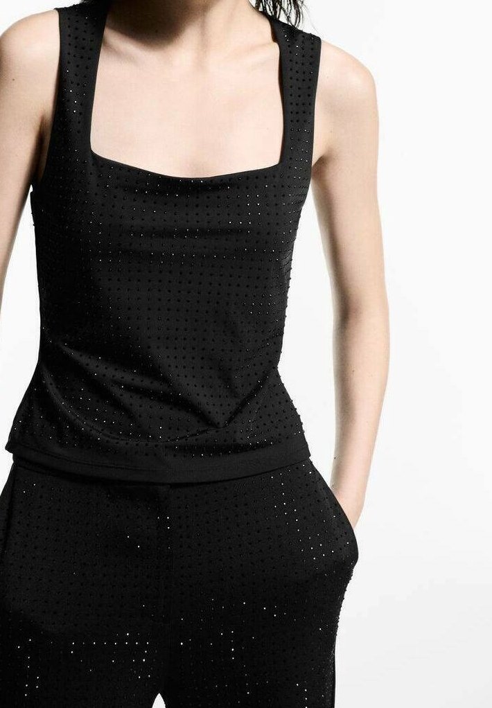 Femme portant un haut noir sans manches et un pantalon assorti avec de petits trous perforés, debout avec une main dans la poche, sur fond blanc.