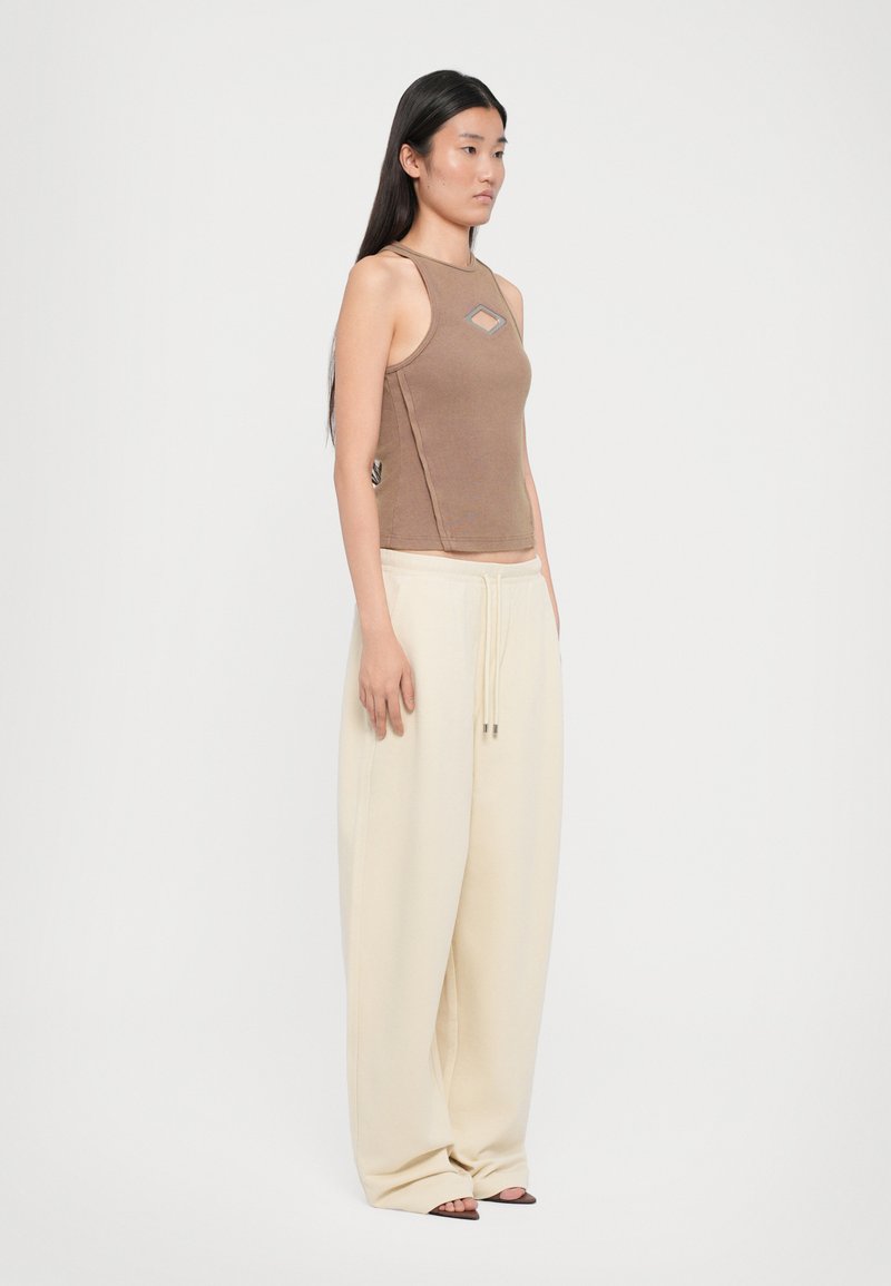 Brown ribbed sleeveless top with a diamond cutout, paired with loose cream-colored trousers made of soft fabric. Simple sandals complete the look.