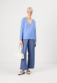Un pull-over bleu clair à col en V et un pantalon bleu ample, associés à un sac à main blanc et des chaussures beiges avec des boucles, sur fond blanc.