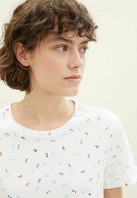 T-shirt blanc avec un col rond, présentant un motif éparpillé de designs en coups de pinceau bleu, marron et noir. Manches courtes, en tissu doux.