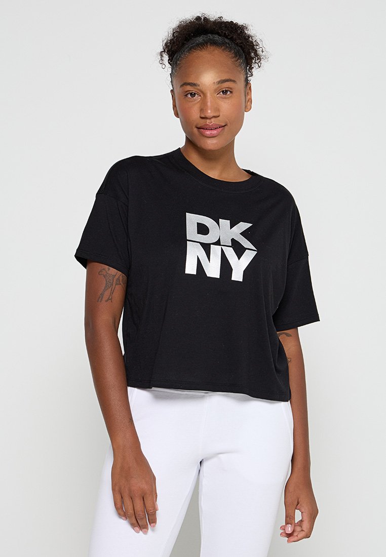 Femme avec des cheveux bouclés portant un T-shirt noir court avec le logo argenté "DKNY" et un pantalon blanc, se tenant contre un fond clair.