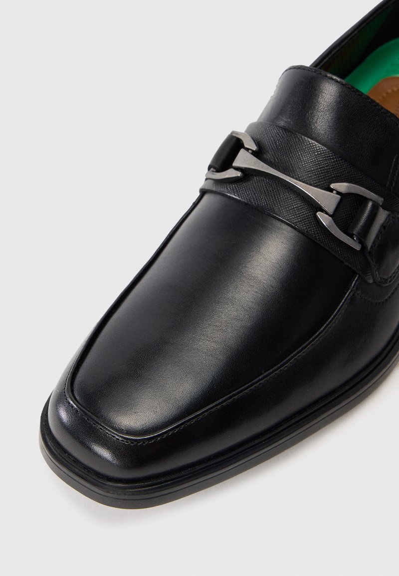 Zwarte leren loafers met een vierkante neus, voorzien van een textuurstrip en metalen accent op de bovenkant. Glad oppervlak en groene binnenzool.