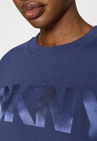 Tmavě modrá mikina z bavlněné směsi, s velkým, výrazným tmavě šedým logem "DKNY" s texturovaným povrchem. Viditelný detail kulatého výstřihu.