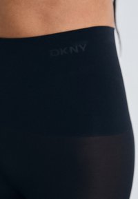 Colanți negri cu talie înaltă, cu o textură fină, având un logo discret DKNY pe talie și o secțiune translucidă în partea inferioară a piciorului.
