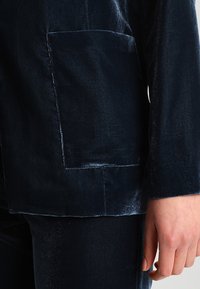 Detailní záběr na osobu v tmavě modrých sametových kalhotách a saku se čtvercovou kapsou, ruka uvolněně položená vedle těla.