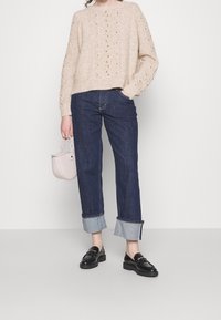 Beige gebreide trui met textuurpatronen, gecombineerd met donkerblauwe spijkerbroek met opgerolde zomen en zwarte loafers. Houdt een kleine beige tas vast.