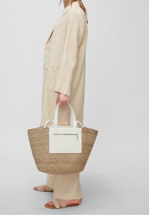 Kvinna klädd i en beige linnedräkt och sandaler som håller en stor vävd stråväska med vit läderficka och handtag.