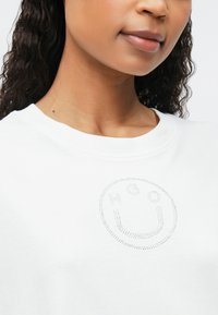 Detailný záber na osobu v bielej košeli s malým dizajnom usmievajúcej sa tváre s bodkami na hrudi.