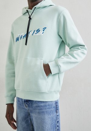 Man in lichtgroene hoodie met voorzak en zwarte trekkoorden, draagt een blauwe spijkerbroek, tekst op de hoodie gedeeltelijk zichtbaar met de woorden "WHAT IS?"