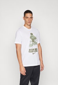 Bela bomba iz bombaž, ki ima zeleno grafiko s tekstom "PREKINI VEČ KOT POT" in Nike logotipom. Kroglično izrezovanje in kratki rokavi.