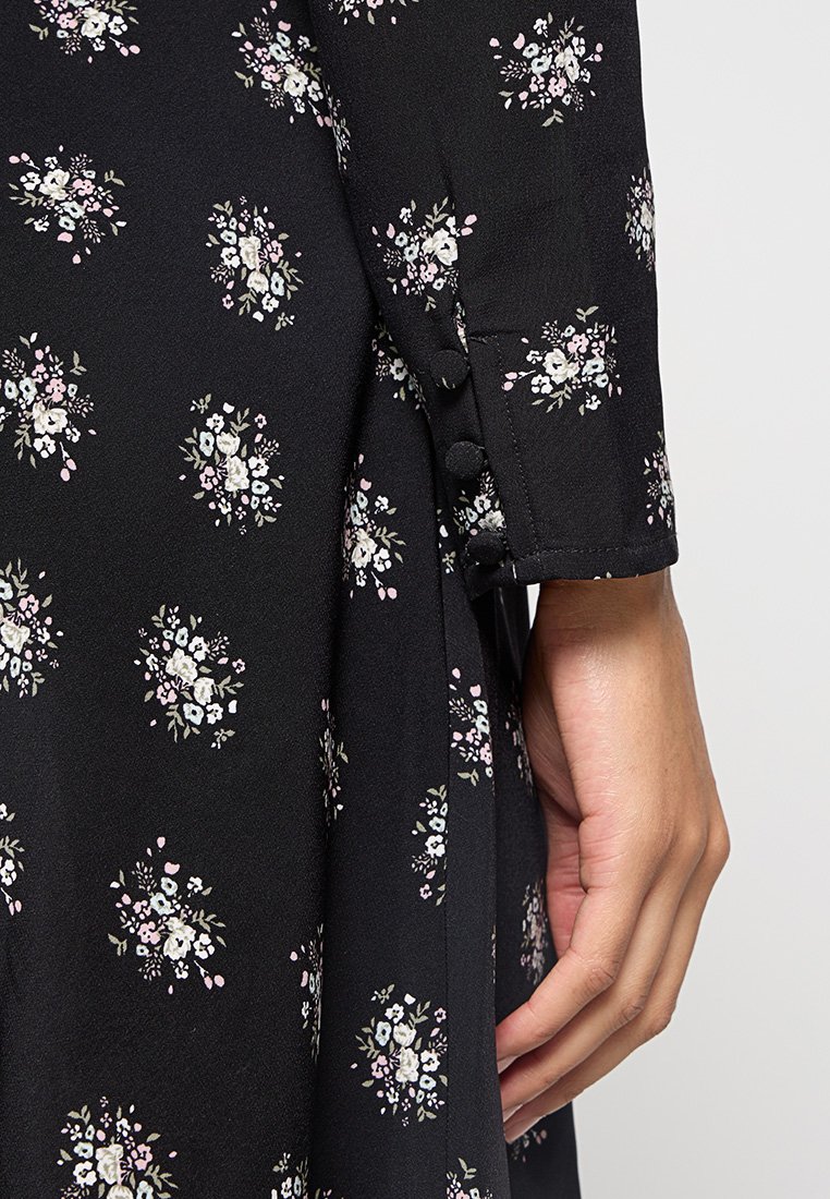 Vestito nero floreale con piccoli fiori bianchi, rosa e blu. Presenta bottoni sulle maniche e una texture del tessuto liscia e morbida.