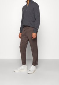 Grauer Zip-Hals-Pullover, braune maßgeschneiderte Hose und weiße Sneaker. Die Hose hat eine glatte Textur; der Pullover ist gestrickt und figurbetont.