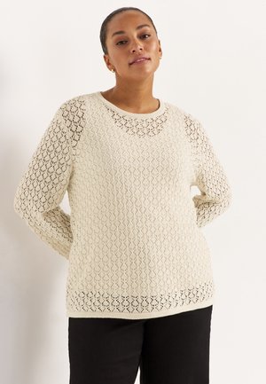 Vrouw draagt een crèmegekleurde, lange mouw gehaakte trui en zwarte broek, staand met handen achter haar rug tegen een witte achtergrond.