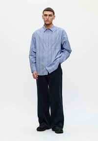 Ung mann iført en løs blåstripet skjorte med knapper, svarte vide bukser og svarte sko, står mot en enkel hvit bakgrunn.