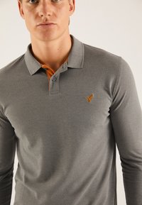 Tricou polo gri cu mâneci lungi și țesătură texturată, având un accent portocaliu sub guler și un mic logo brodat pe piept.