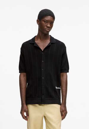 Mustast lühikeste varrukatega särk, millel on tekstureeritud kudumismuster, vertikaalsed triibud, kraega kaelus ning valge logo detail allääril.