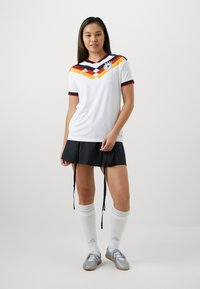 Jeune femme debout, portant un maillot de football blanc de l'Allemagne, une jupe noire, des chaussettes blanches montantes jusqu'aux genoux et des crampons argentés, sur un fond uni.