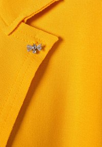 Tessuto giallo con una texture liscia, caratterizzato da una spilla a forma di fiocco in argento con una perla e un piccolo accento di pietra preziosa vicino al colletto.