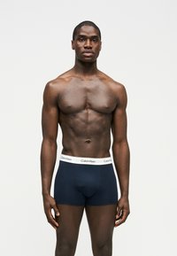 Hombre musculoso de pie, mirando hacia adelante, con bóxer Calvin Klein azul marino y cinturilla blanca, sobre un fondo liso.