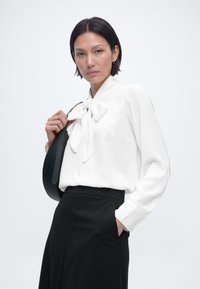 Γυναίκα με λευκό μπλουζάκι μακρυμάνικο με φιόγκο μπροστά, μαύρη φούστα ψηλόμεση, κρατώντας μαύρη τσάντα ώμου, στέκεται μπροστά σε απλό ανοιχτό φόντο.