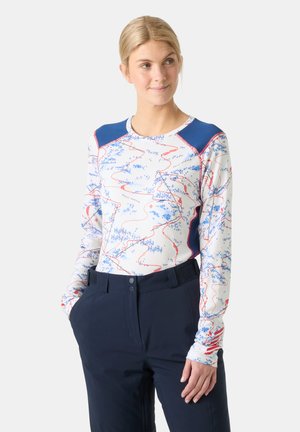 Mujer con cabello rubio que lleva una camiseta blanca de manga larga con líneas abstractas azules y rojas y pantalones azul marino, de pie con las manos en los bolsillos.