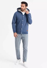 Sudadera azul con botones y un interior claro en contraste. Cuenta con un bolsillo tipo canguro, puños acanalados y una etiqueta de acento de cuero. Combinada con jeans claros y zapatillas.