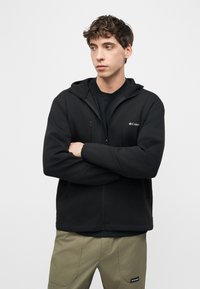 Νεαρός άνδρας με σγουρά μαλλιά, φορώντας μαύρο φούτερ Columbia με φερμουάρ και χακί παντελόνι, στέκεται με σταυρωμένα χέρια, κοιτάζοντας πλάγια.