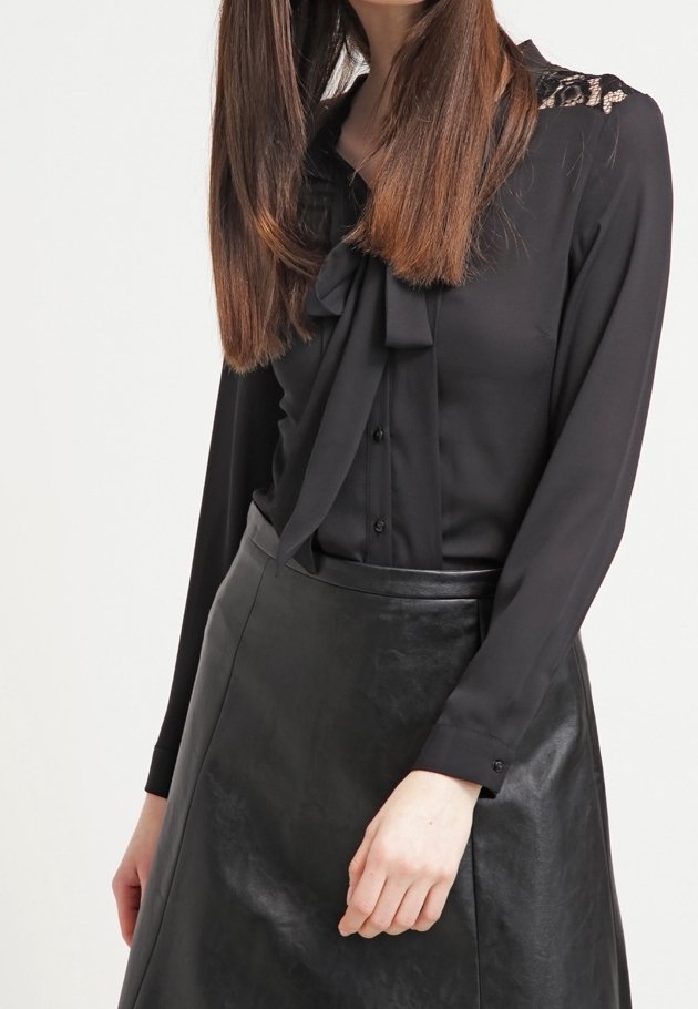 Blusa nera con fiocco, apertura frontale a bottoni e dettagli in pizzo sulle spalle, abbinata a una gonna in pelle nera, dalla texture liscia.