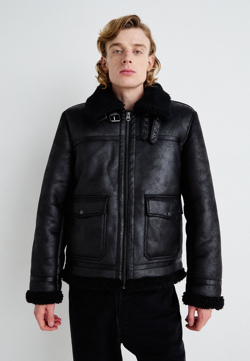 Black leather jacket with a shearling collar, front zipper, snap button pockets, and textured finish. Accented with buckle details on the collar.