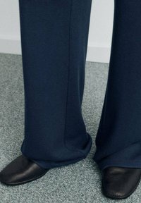 Pantalon bleu marine foncé couvrant légèrement des chaussures noires à bout carré sur un sol moquetté gris.