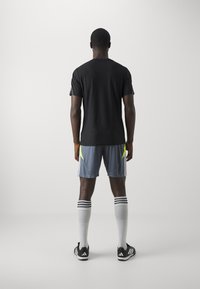 Svart kortermet skjorte, grå shorts med gule detaljer, hvite knelengde sokker og svarte sportssko. Bakgrunnsutsikt mot en ensfarget bakgrunn.