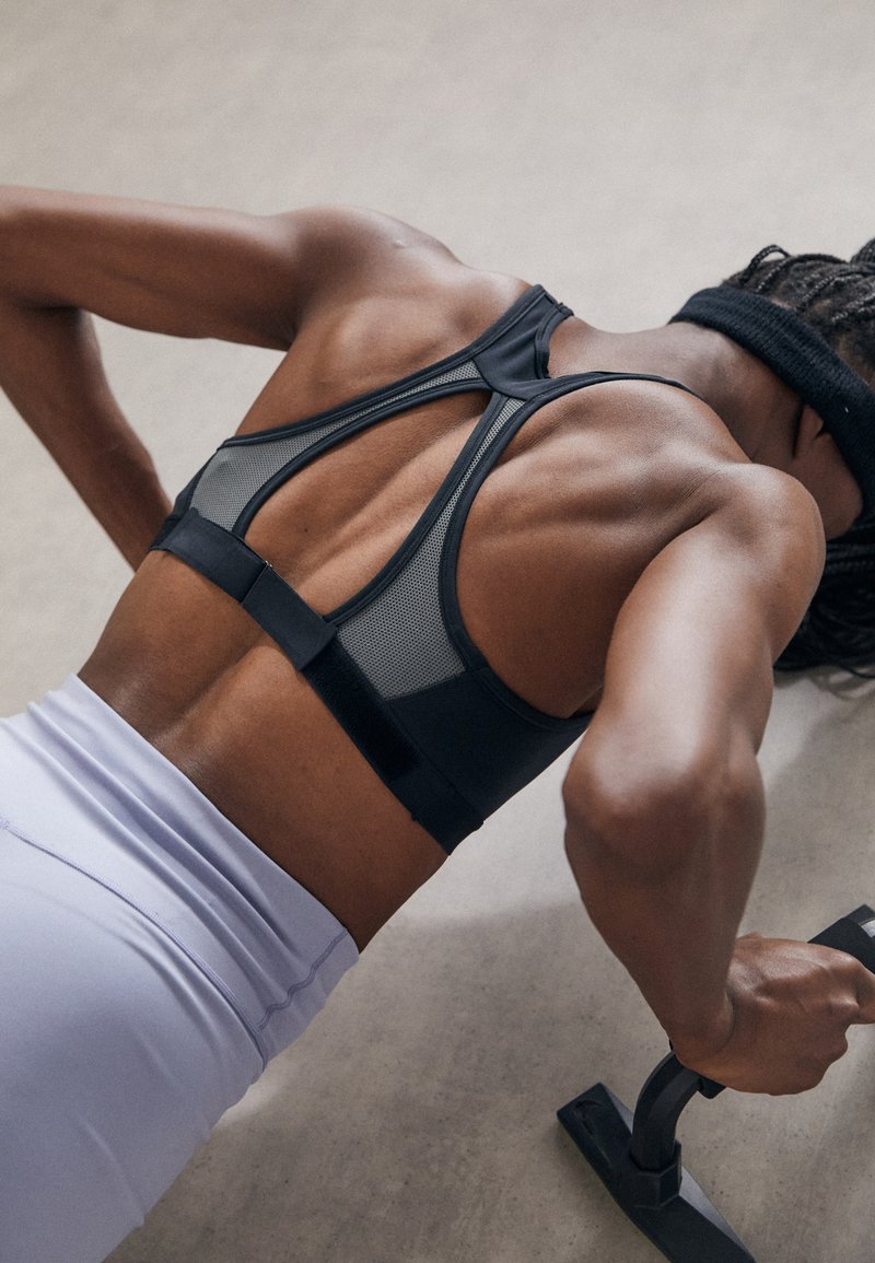Black mesh sports bra with a racerback design, paired with light purple workout pants. Focus on athletic posture and grip on push-up handles.