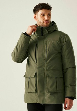 Hombre con cabello corto y rizado que lleva una chaqueta de invierno verde oliva con múltiples bolsillos con cremallera y solapa, ajustándose el cuello.